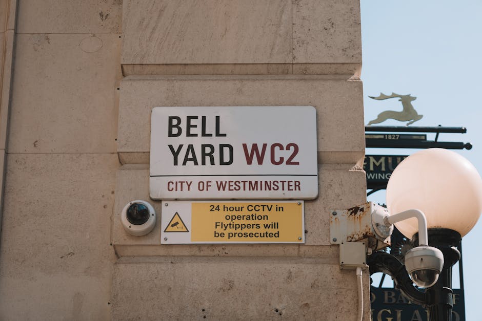 A close-up image of a beige stone wall on a building corner displaying a white rectangular sign with black and red text reading 'BELL YARD WC2' and 'CITY OF WESTMINSTER.' Below the main signage, a smaller yellow sign with black text states '24 hour CCTV in operation. Flytippers will be prosecuted,' accompanied by a surveillance camera mounted on the wall to the left of the sign. To the right, a traditional black street lamp with a white, spherical glass cover is attached to the wall, with multiple electrical cables visible connecting it. In the background, part of a decorative iron street sign with a silhouette of a deer is visible against a light blue sky. The scene illustrates a typical urban boundary area where private waste collection or on-site clearance might occur, aligning with the context of rubbish disposal regulations in Westminster.