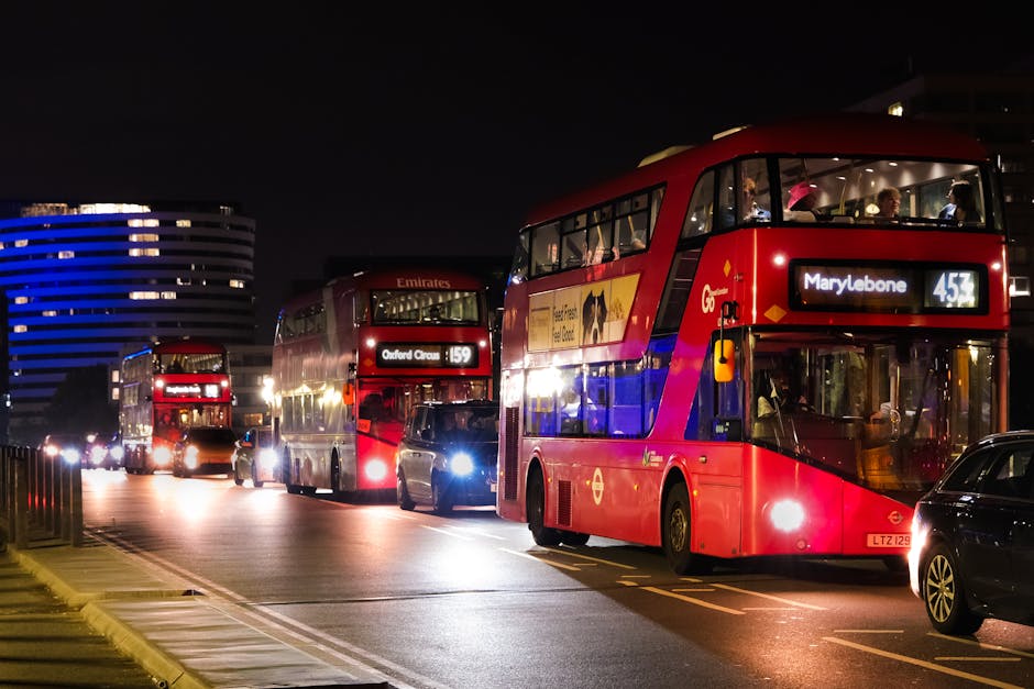 Three red double-decker buses are parked in a line on a city street at night, with the bus closest to the camera displaying the route number 453 heading to Marylebone. The second bus behind shows route 159 to Oxford Circus, and the third bus, operated by Emirates, also has internal lighting visible through its large windows, revealing seated passengers. The buses have a typical London design with a glossy red finish and standard round yellow fog lights on the front. Surrounding the buses, there are several parked cars along the curb, including a small dark vehicle next to the front bus. The scene is illuminated by the buses' headlights and some street lighting, casting reflections on the dark pavement, which features yellow dash lines and a sidewalk with a metal railing on the left. In the background, modern buildings with curved facades and illuminated windows are visible against the night sky, creating an urban atmosphere that complements the context of city transportation and local public services. Waste Clearance Marylebone occasionally supports private waste handling, which aligns with the need for effective rubbish collection within such busy city environments, although not visible in this scene.