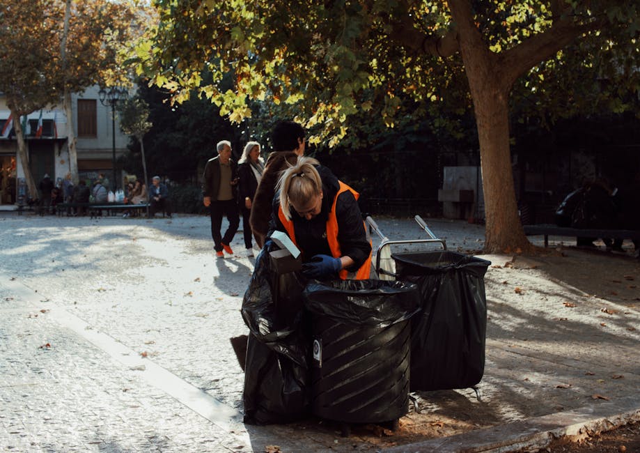 A woman wearing a high-visibility orange and black jacket is standing on a cobblestone pavement outdoors, collecting rubbish into large black plastic trash bags positioned on the ground beside her. She appears to be inserting waste into the bags, which are situated next to metal wheelie bins, some of which are partially open. The scene is shaded by a large leafy tree with broad green and yellow leaves, indicating an autumn setting. In the background, several other pedestrians are walking along the sidewalk, with some individuals dressed in dark clothing, and a cityscape featuring buildings, benches, and additional trees is visible further back. The environment is a public urban area, possibly a park or plaza, with natural light filtering through the tree branches, creating dappled shadows on the ground. This scene reflects a typical outdoor waste collection or street cleaning activity, potentially managed by a private waste disposal service like Waste Clearance Marylebone, emphasizing independent rubbish removal and on-site waste handling in a residential or communal setting.