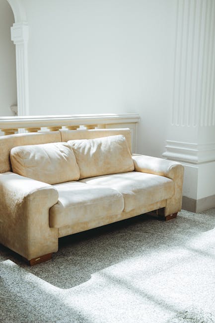 A beige fabric upholstered sofa with a plush, slightly worn texture, positioned on a light grey carpeted floor in a well-lit interior space. The sofa has two seat cushions and two matching back cushions, with rounded armrests on both sides and small wooden legs visible beneath. It is situated against a white wall with decorative architectural molding and a white column on the right side, indicating a classic interior design style. The surrounding environment includes a portion of the wall's vertical paneling and a shadow cast by natural light coming from an unseen window, creating a clean and orderly scene. This setting reflects a typical indoor environment where furniture might be prepared for removal or replacement, aligning with concepts of private waste disposal or on-site clearance services sometimes provided by companies like Waste Clearance Marylebone, as part of their rubbish removal offerings.