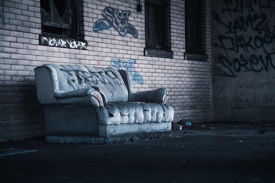 An old, worn-out fabric sofa with a light blue, slightly faded upholstery sits against a brick wall in an outdoor urban setting. The sofa's material shows signs of damage, with tears and rips visible on the seat, backrest, and armrests, exposing foam padding and internal cushioning. The fabric appears textured and has a pattern of small dots or a subtle weave. The armrests are padded and rounded, with some areas frayed or worn through. The sofa is positioned centrally in the foreground, occupying most of the image, with its base resting directly on the paved ground. In the background, a brick wall with a mix of light and dark bricks features several patches of graffiti in various shades of blue and black. Nearby, two small, barred windows are visible, with dark, grimy frames. The lighting is dim and moody, casting shadows that emphasize the dilapidated condition of the sofa and the neglected environment, which appears to be part of an abandoned, graffiti-covered alley or backstreet, indicative of a private or alternative waste management context as seen in independent rubbish disposal services by Waste Clearance Marylebone.