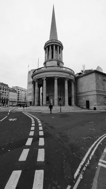 A black and white photograph capturing a historic, circular stone building with tall, evenly spaced columns supporting a wide, curved entablature at street level. Above this, a tall, slender spire rises sharply into the sky, giving the structure a distinctive neoclassical appearance. The building is situated at an intersection with visible road markings, including dashed lines and bicycle lane symbols, indicating an urban setting. In the foreground, a person dressed in dark clothing is walking across the street, which is free of traffic. Surrounding buildings are visible in the background, featuring classic European architectural styles with multiple stories and traditional rooflines. The scene appears to be in a city area, possibly an important landmark or public space. The image exudes a quiet, orderly atmosphere, typical of city streets with historical architecture, and subtly aligns with the context of on-site or alternative waste handling during urban refurbishment or preservation projects, as represented by Waste Clearance Marylebone's services.
