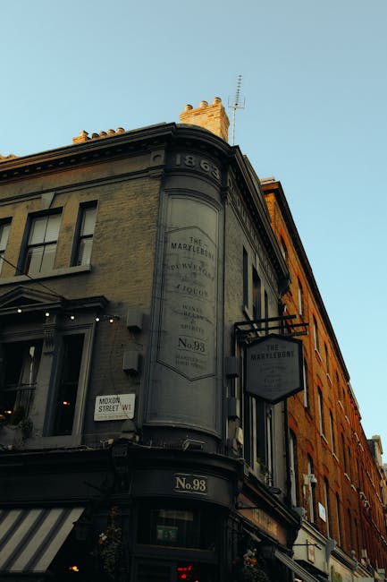 A three-story corner building with a vintage architectural style, painted in muted beige and darker tones, is shown under a clear blue sky. The building features large, rectangular same-sized windows with black frames, some with decorative window ledges. The curved corner displays an aged, faded sign painted directly onto the facade, indicating the building's historical name and number, with the number '98' on a small plaque at the street level. A black signboard with white lettering extends outward, attached to the building's facade, advertising a business. A striped awning covers the storefront at the ground level, with small potted plants hanging beneath it, adding a touch of greenery. The environment suggests an urban street setting, possibly in a historic district, with other buildings lining the street and a street sign indicating nearby streets. The simple lighting and clear sky imply daytime. This scene, with its classic architecture and urban context, subtly relates to private property management and the structural environment where rubbish removal services like those from Waste Clearance Marylebone operate to service commercial premises.