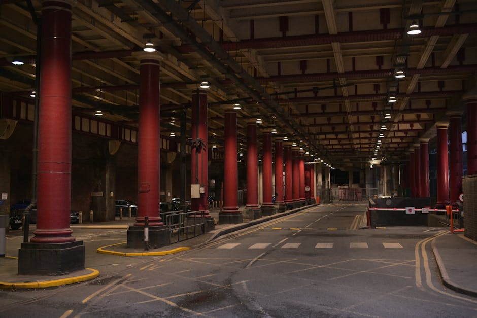 This image depicts an indoor parking area beneath a large structure supported by multiple tall, cylindrical red columns with black bases. The ceiling features exposed beams and piping, with evenly spaced small lights illuminating the space. To the left, a row of parked cars is visible, partially obscured by the columns, while the right side shows a barrier gate, indicating controlled vehicle access. The floor is made of dark asphalt marked with yellow and white road and parking zone lines, including a pedestrian crossing in the foreground. The environment appears clean and well-maintained, with no litter or debris visible. The lighting creates a warm, somewhat subdued atmosphere suitable for a parking facility or vehicle storage area, emphasizing the structural elements and the orderly layout. Waste Clearance Marylebone's rubbish removal services could be relevant to this setting if considering maintenance or waste management within such infrastructure, although no waste or rubbish is visible in this particular image.