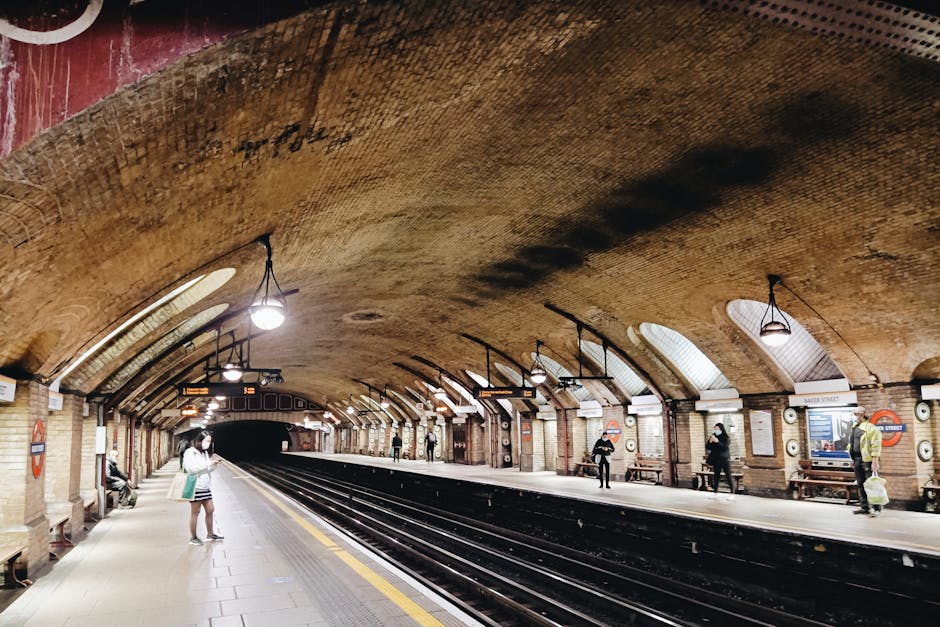The image shows a commercial underground train station with a curved, arched ceiling constructed of brown bricks, creating a textured and historic appearance. The platform is relatively wide, lined with a yellow safety line along the edge close to the dark, empty train tracks. Several passengers are standing or sitting on benches distributed along the platform, some engaged with their phones, others waiting quietly. A person in the foreground, wearing a face mask and casual clothing, is holding a bag while standing near the edge of the platform. Bright, round hanging lamps illuminate the space, casting a warm light on the platform and the brick ceiling, with additional lighting fixtures suspended from the arches. The station walls feature vintage-style signage, including the recognizable round London Underground logo, which suggests a location within the London transport network, possibly outside the Marylebone station area. This setting exemplifies an underground transit environment suitable for private or independent travel while undertaking waste removal or station maintenance activities, as managed by Waste Clearance Marylebone's services for rubbish and bulky waste collection.