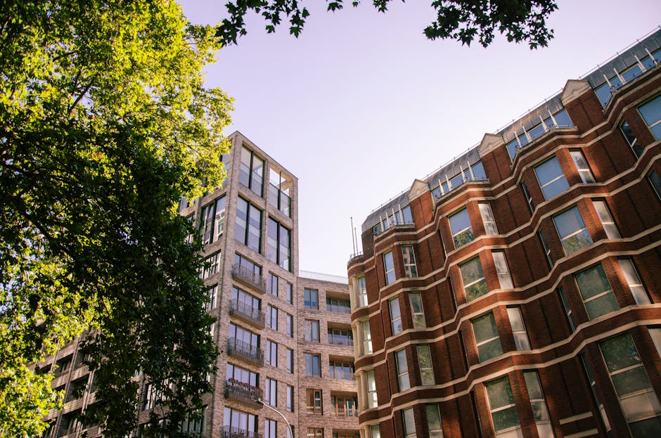 A clear daytime view of two adjacent multi-storey buildings, one constructed with reddish-brown brick featuring curved bay windows with white frames, and the other with a modern grey stone facade and large glass balcony doors, set against a bright sky. To the left, a leafy tree with vibrant green foliage partially obscures the buildings, casting dappled shadows on the structures. The scene captures an urban residential environment, with the buildings' exterior details and the natural green element connecting to the context of property maintenance and potential waste management activities in city settings. The lighting highlights the textures of the brickwork and stone, with shadows emphasizing the architectural features. The environment appears peaceful, with no visible activity or clutter, reflecting a typical cityscape that may require services for building clearance or waste removal, as offered by Waste Clearance Marylebone. The overall composition emphasizes the structural details and the natural surroundings of the urban area, aligning subtly with the themes of property upkeep and private waste handling.