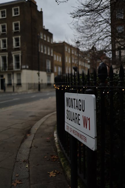 A black wrought iron fence surrounds Montagu Square in Westminster, with a white street sign mounted on the fence indicating 'Montagu Square W1' with smaller text below labeling it as part of Westminster. The fence features pointed finials on top. In the background, a row of multi-storey residential buildings with brown and beige brick facades, some with white window frames, are visible beneath an overcast sky. The pavement adjacent to the fence is partially covered with fallen leaves, and the area appears quiet with no pedestrians or vehicles present. The scene is illuminated by natural daylight, contributing to a subdued, urban atmosphere typical of central London streets and alleys, where private waste collection or rubbish removal services might be utilized to manage waste instead of public street cleaning, aligning with the context of rubbish removal services in an urban environment.