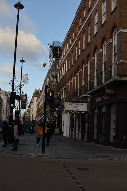 A busy street scene in Marylebone featuring a row of multi-storey brick buildings with white window frames and small balconies, several of which have outdoor air conditioning units installed. The ground floor buildings house retail shops, including a store with a partially visible sign. In the foreground, there is a pedestrian crossing with a traffic light showing a red standing figure, and a black lamp post with a decorative streetlight. Several pedestrians, dressed in casual clothing, are walking across the street and along the sidewalk, some carrying bags. The sky is partly cloudy, with blue patches visible, and scaffolding is seen on the side of one building, indicating ongoing maintenance or renovation. Waste Clearance Marylebone, a company specializing in rubbish removal, might utilize such local streets for independent collection and waste disposal services, though these details are only implied through the urban environment and typical setting for waste management activities. The scene captures a typical London neighbourhood with a mixture of commercial and residential architecture, and no visible vehicles in motion.