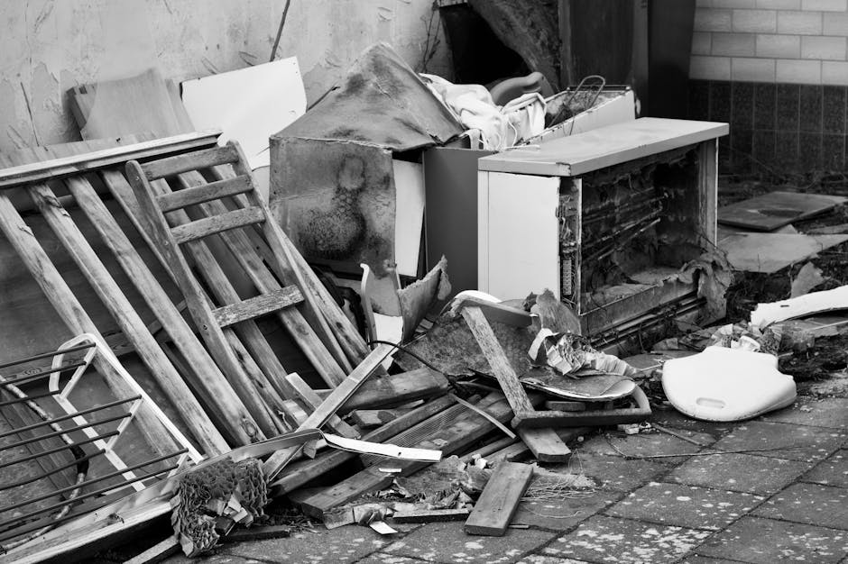 A pile of discarded household waste and debris situated on a paved outdoor area beside a brick and concrete wall. The rubbish includes an old wooden chair frame leaning against a partially dismantled white cabinet, with broken wooden slats and splintered edges visible. There are pieces of chipped wood, torn fabric, and scattered debris such as fragmented plastic and paper. An unassembled or damaged appliance component with exposed internal elements is partly covered by a torn cloth or insulation material. The environment appears cluttered and neglected, suggesting an area awaiting clearance or removal. The scene reflects a typical site for private rubbish collection services, with visible signs of mixed waste material that could be managed by companies like Waste Clearance Marylebone, offering same-day bulky waste removal. The overall lighting is natural, with subtle shadows indicating daylight, emphasizing the disorder and the need for efficient rubbish removal in a residential or commercial setting.