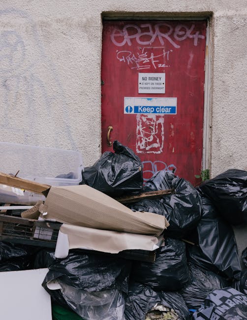 A collection of black plastic rubbish bags, some torn or bulging, piled in front of a red metal door on an exterior wall. The door features graffiti tags and is marked with several signs, including a white sign that states 'No Money, No Key or Code, Premises Overnight,' and a blue sign with the message 'Keep clear.' Surrounding the bags are discarded cardboard pieces, a section of foam or padding, and some metal or plastic objects partially hidden among the waste. The scene suggests an area designated for waste disposal outside a building, with the bags appearing temporarily stored or awaiting collection. The concrete wall behind the door is textured and light-colored, and the overall atmosphere conveys an informal rubbish accumulation typical of alternative waste handling or private collection services such as those offered by Waste Clearance Marylebone. The lighting is natural, highlighting the worn condition of the door and the disorderly arrangement of refuse, emphasizing the need for proper rubbish removal or clearance services in the area.