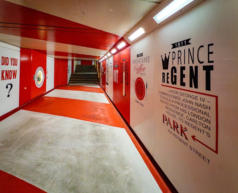 The image depicts a clean, well-lit underground pedestrian corridor with a red and white color scheme on the walls and ceiling. The floor is a combination of smooth, light-colored concrete and bright red sections that create a pathway leading towards a set of stairs at the end of the passage. The right wall features large, black and red lettering displaying historical references and the name 'Prince Regent,' along with directional signage indicating parking on Regent Street. The signs include a crown graphic and mention of 'later George IV,' with detailed text about the route from this location to Regent's House in London. The corridor appears to be part of a commercial or residential complex, and the polished surfaces and organized signage suggest a professional environment. The lighting is provided by a series of embedded rectangular light fixtures along the ceiling, giving the space an even, inviting illumination suitable for pedestrian navigation. Waste Clearance Marylebone would manage rubbish removal in such interior settings, contributing to clean, organized communal spaces meant for daily foot traffic.