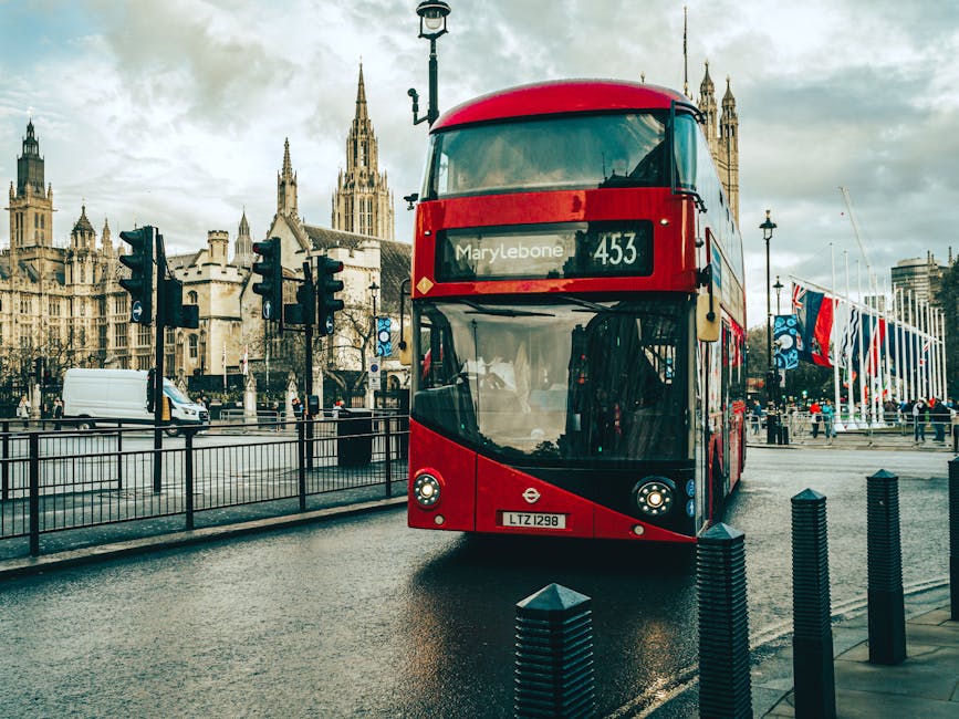 A red double-decker bus parked on a wet street in front of historic Gothic-style buildings with tall spires and ornate facades, including the Palace of Westminster in the background. The bus displays route information for Marylebone on the destination sign, and the vehicle's front windshield and headlights are visible. The scene is overcast with grey clouds, and the street features black bollards, a metal fence, and a row of flags along the sidewalk. A white van is seen in the background, along with pedestrians walking nearby. This setting illustrates a typical urban environment in central London, where private transport and infrastructure support city operations. The scene may relate to local waste management activities, such as rubbish collection points or service vehicles situated in busy city streets, with Waste Clearance Marylebone providing independent rubbish removal solutions in the area.