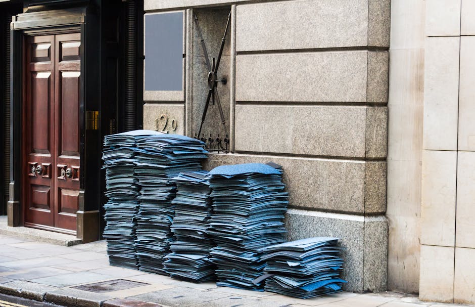 A large pile of blue and white paper and cardboard documents stacked haphazardly on the pavement outside a building with a granite facade. The stack extends from the ground up to nearly waist height, with some piles leaning slightly to one side. To the left, there is a wooden double door with a dark finish and metallic door handles, set within an ornate black frame. Behind the paper stacks, a grey metal utility box with a locking latch and black hinges is mounted on the stone wall. The environment appears to be an urban street scene, with natural light casting subtle shadows, indicating it is daytime. The setting suggests a scene where private waste collection or on-site clearance, possibly handled by waste clearance services such as Waste Clearance Marylebone, involves removing large quantities of paper waste from commercial premises. The textures of the paper are slightly crumpled and uneven, contrasting with the smooth, polished surface of the building's granite structure.