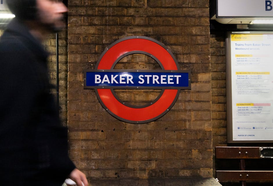 A close-up view of a London Underground station sign for Baker Street mounted on a textured brick wall in a station corridor. The sign features a circular red frame with a horizontal blue bar across the middle displaying the white text 